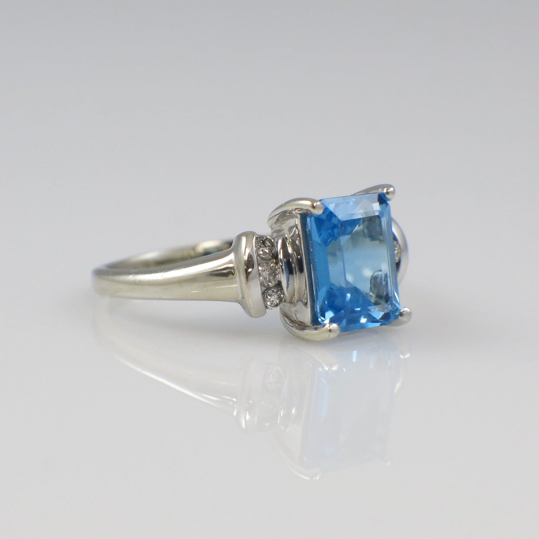 14K white gold blue topaz ring with diamond accents, emerald cut, featuring a sparkling blue gemstone and intricate detail.